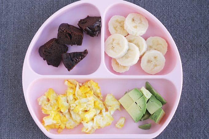 A pink plate with sections containing different foods such as avocado and bananas.