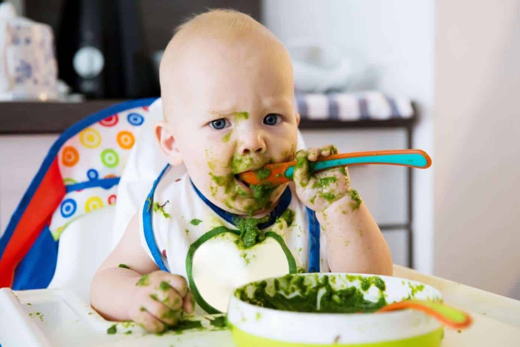 A toddler eating greens from a aplate with a spoon in a messy way
