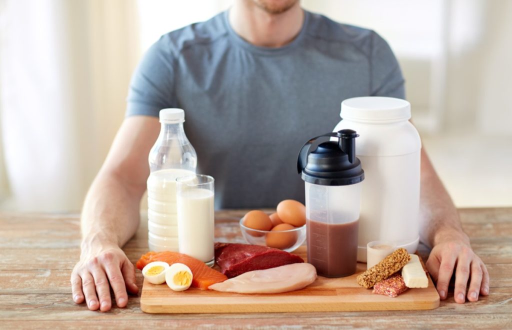 A man on a table with foods rich in proteins such as milk, stealk, eggs and protein shakes.