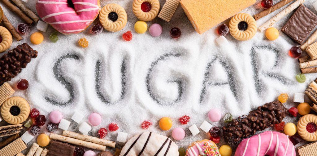 Sugar poured on a table and the word sugar written on it. On the sides are candy, cakes, chocolate, biscuits and other items containing sugar.