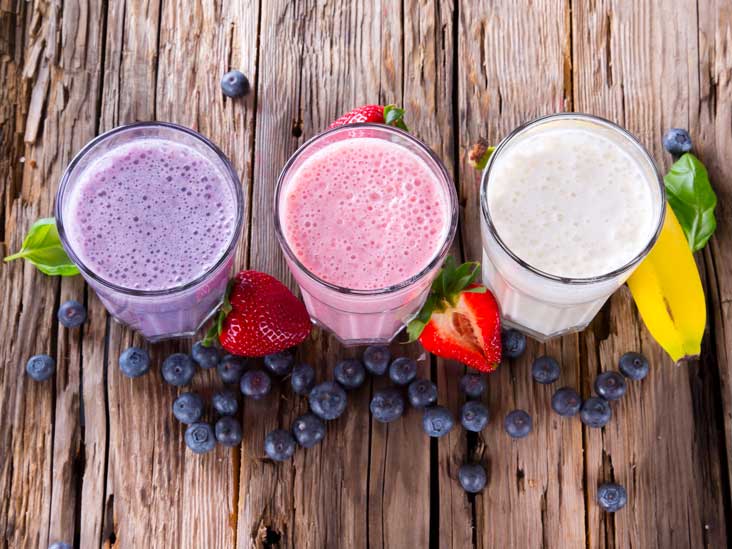 Three glasses of protein shakes on a table with grapes, straw berries and a banana.
